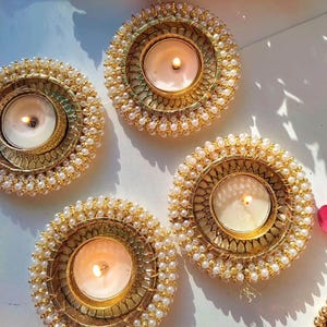 May include: Four circular candle holders featuring a gold-toned metal frame with a decorative lattice pattern. Each holder is adorned with a border of small white faux pearls and contains a single lit white tea light candle in the center.