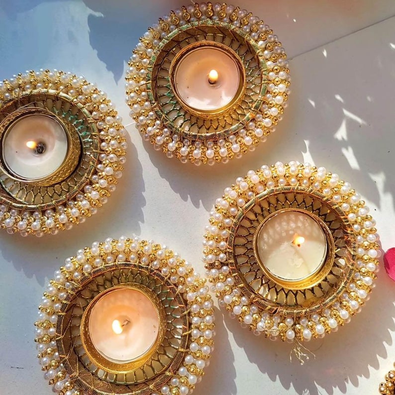 May include: Four circular candle holders featuring a gold-toned metal frame with a decorative lattice pattern. Each holder is adorned with a border of small white faux pearls and contains a single lit white tea light candle in the center.