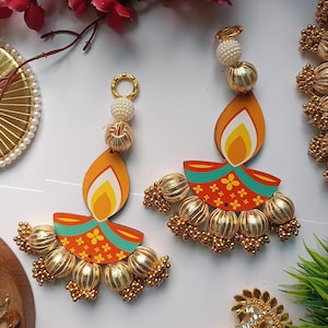 May include: Two decorative earrings in the shape of a diya, a traditional oil lamp. The earrings feature a flame design in orange, yellow, and white, with a red base adorned with floral patterns. Gold-colored beads and accents complete the design.