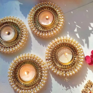 May include: Four circular candle holders featuring gold-toned metal frames with intricate lattice patterns and borders lined with small white faux pearls. Each holder contains a lit white tea light candle, casting a warm glow on the surface.
