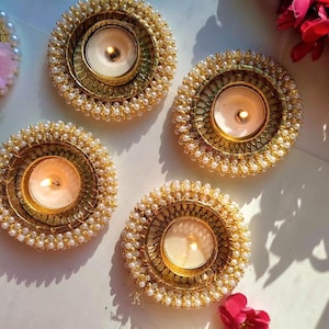 May include: Four circular candle holders featuring a metallic gold finish with intricate textured patterns and a border of small white beads. Each holder contains a lit tea light candle with a warm yellow flame, creating a soft glow.