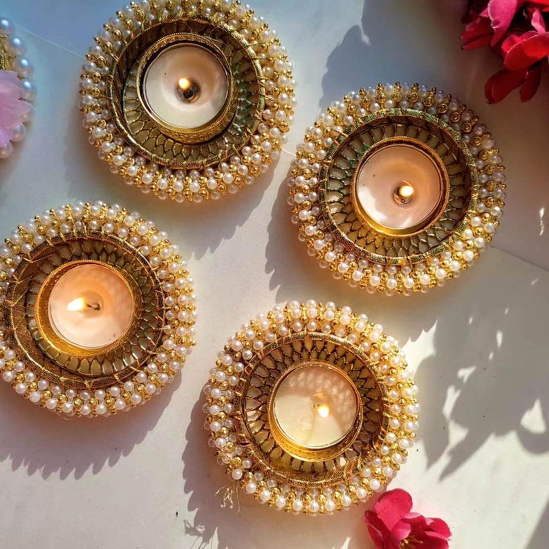 May include: Four circular candle holders featuring a metallic gold finish with intricate textured patterns and a border of small white beads. Each holder contains a lit tea light candle with a warm yellow flame, creating a soft glow.