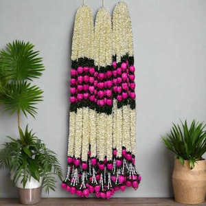 May include: Three white flower garlands with pink flower accents hanging on a wall. The garlands are made of artificial flowers and are used for traditional Indian weddings.