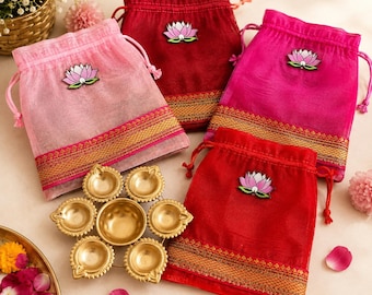 May include: Four drawstring pouches in light pink, dark red, magenta, and bright red, each featuring a lotus flower emblem and a gold-toned border. A gold-colored metal diya lamp with six small bowls arranged in a circular flower pattern.
