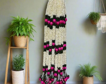 May include: A long, vertical garland with white and dark green sections, punctuated by bright pink flower clusters. The garland is suspended against a light green wall, with potted plants on a wooden shelf and hanging from the ceiling.