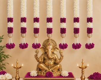 May include: A gold-toned Ganesha statue sits on a rectangular platform with a purple velvet top. Seven vertical floral garlands in white and magenta hang from a gold rod above. Two brass oil lamps and bowls of flowers flank the central platform.
