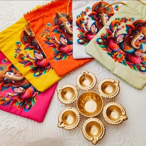 May include: A selection of brightly coloured drawstring bags, each displaying a detailed illustration of a deity. The bags are in pink, yellow, orange, white, and green. A gold-coloured metal oil lamp holder with seven compartments is also visible.