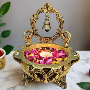 Pu&ograve; includere: Un supporto decorativo in metallo dorato con una ciotola rotonda, intagli intricati e una piccola campana appesa in cima. La ciotola &egrave; piena di acqua e petali di rosa, e una piccola candela brucia al centro.