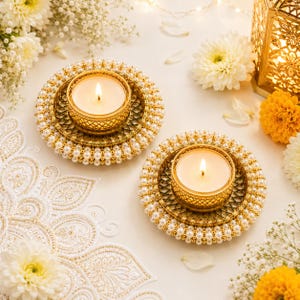 May include: Two circular gold-toned candle holders featuring a central tea light cup surrounded by a decorative lattice pattern and an outer border of small white pearls. Each holder contains a lit white tea light candle with a visible flame.