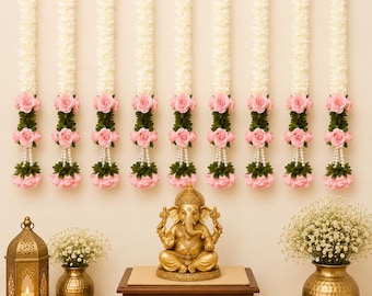 May include: Nine vertical floral garlands featuring white blossoms, pink roses, and green leaves hang above a gold-toned Ganesha statue on a wooden pedestal, flanked by a metal lantern and a vase filled with white baby's breath flowers.