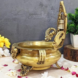 Pu&ograve; includere: Un bruciatore di incenso in ottone dorato con dettagli intricati e un coperchio incernierato. Il bruciatore ha tre gambe e un manico decorativo. &Egrave; seduto su una superficie marrone con petali di fiori sparsi intorno.