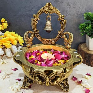 Pu&ograve; includere: Un supporto decorativo in metallo dorato con una ciotola rotonda piena di acqua e petali di fiori. Una piccola candela si trova al centro della ciotola. Una campana pende dalla parte superiore del supporto.