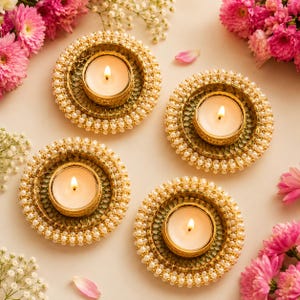 May include: Four circular candle holders featuring a metallic gold finish with intricate lattice patterns and borders lined with small white pearls. Each holder contains a single lit tea light candle with a warm yellow flame, arranged symmetrically.