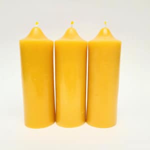 May include: Three cylindrical yellow beeswax candles standing upright in a row. Each candle features a smooth, solid surface with a rounded top and a small, centred wick protruding from the apex. The candles are uniform in colour and shape.