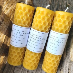 May include: Three cylindrical beeswax candles with a honeycomb pattern and white labels that read "Natural Beeswax Candles 100% Pure Maison Rustique". The candles are yellow and have a wick. They are in a woven basket.