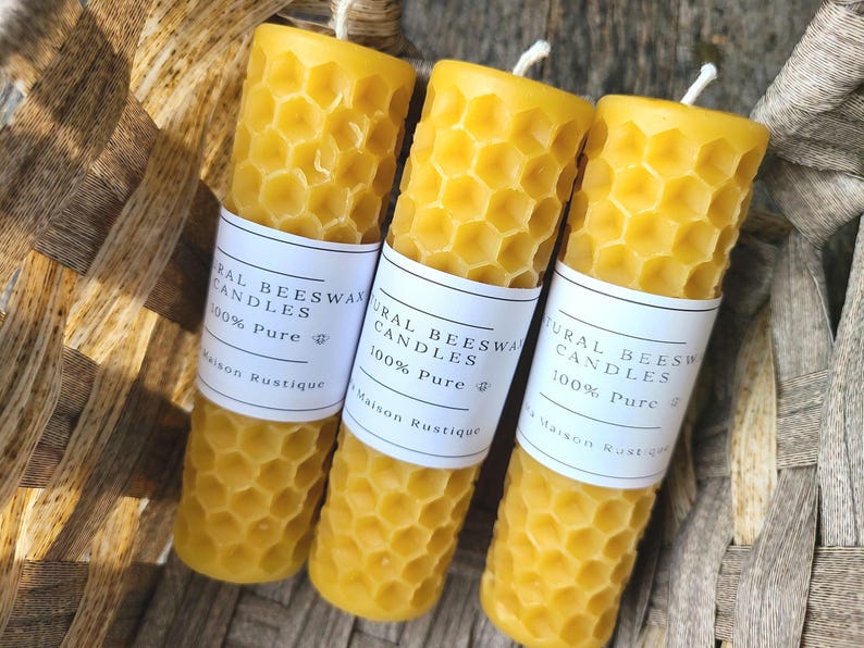 May include: Three cylindrical beeswax candles with a honeycomb pattern and white labels that read "Natural Beeswax Candles 100% Pure Maison Rustique". The candles are yellow and have a wick. They are in a woven basket.