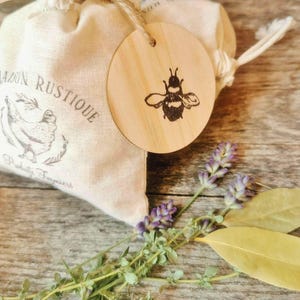 May include: A small, beige linen bag with a drawstring and a wooden tag with a bee illustration. The bag has a printed design with the words "MAISON RUSTIQUE" and a hen illustration. Lavender and bay leaves are arranged nearby.