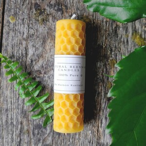 May include: A cylindrical beeswax candle with a honeycomb pattern. The candle is yellow and has a white label that reads "Natural Beeswax Candles 100% Pure". The candle is approximately 4 inches tall. The background is a wooden surface with green foliage.