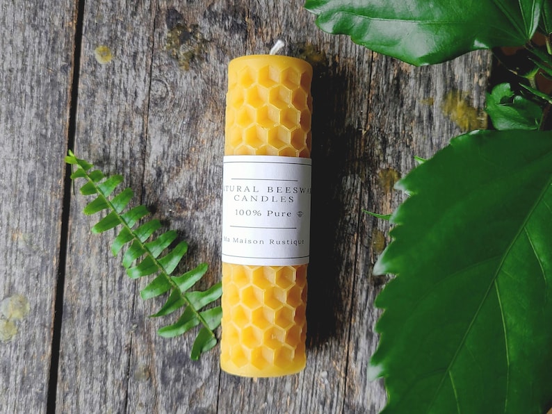 May include: A cylindrical beeswax candle with a honeycomb pattern. The candle is yellow and has a white label that reads "Natural Beeswax Candles 100% Pure". The candle is approximately 4 inches tall. The background is a wooden surface with green foliage.
