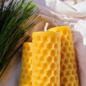 May include: Three yellow beeswax candles with a honeycomb pattern, next to a sprig of green pine needles. The candles have a cylindrical shape and a textured surface. The background is a white fabric.