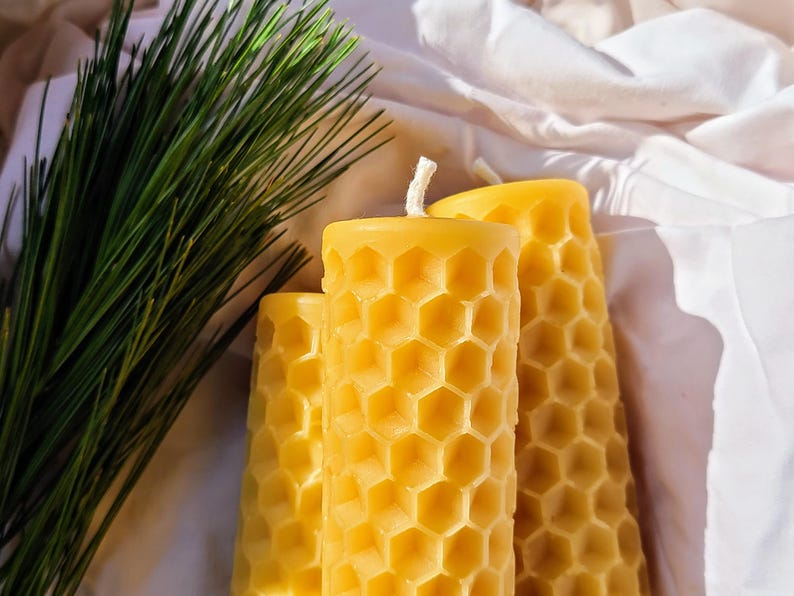 May include: Three yellow beeswax candles with a honeycomb pattern, next to a sprig of green pine needles. The candles have a cylindrical shape and a textured surface. The background is a white fabric.