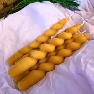 May include: A set of four yellow taper candles with a distinct spiral, twisted design. Each candle features a smooth, matte finish and a single white wick at the top. The candles are arranged in a cluster, showcasing their uniform golden-yellow colour.