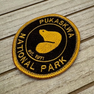 Könnte beinhalten: Ein gestickter Aufnäher in Gold und Schwarz mit dem Text "Pukaskwa National Park est. 1971" und einer stilisierten Biber-Silhouette.