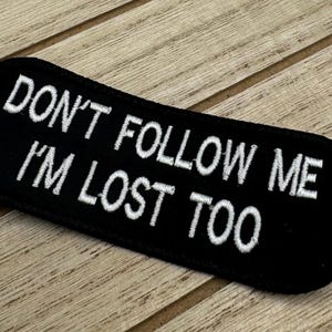 Op de afbeelding: Zwarte geborduurde patch met witte tekst die "Volg me niet, ik ben ook verdwaald" zegt.