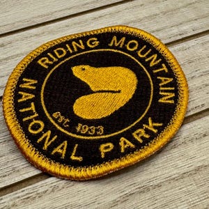 Op de afbeelding: Een gouden geborduurde patch met de woorden "National Riding Mountain Park Est. 1933" in zwarte tekst rond een zwarte cirkel met een gouden beren silhouet erin.