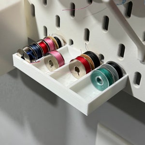 May include: A white plastic spool holder with multiple spools of thread in various colours, including red, pink, blue, black, and green. The holder is attached to a white pegboard.