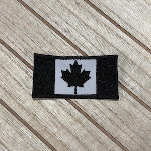 Puede incluir: Un parche rectangular negro y gris con la bandera canadiense. El diseño presenta una hoja de arce negra centrada en un fondo gris, con bordes negros. El parche se muestra sobre una superficie de madera.