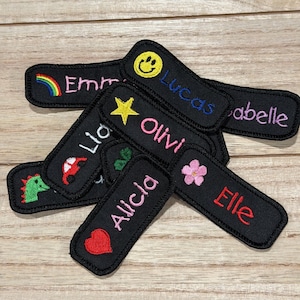 May include: A collection of black embroidered name patches. Each patch features a different name in colorful thread, along with small embroidered icons like a rainbow, star, heart, flower, and a smiley face. The names include Emma, Lucas, Olivia, Alicia, and Elle.