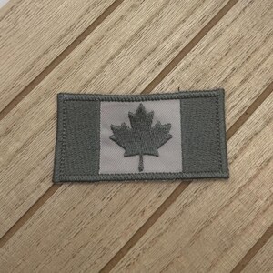 Puede incluir: Un parche bordado gris con la bandera canadiense. La bandera es blanca con una hoja de arce roja.