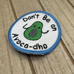 Può includere: Toppa ricamata bianca con bordo blu. La toppa presenta un avocado verde con un viso e il testo "Don't Be An Avoca-dho".