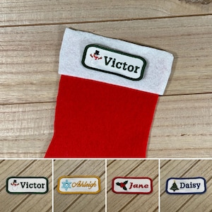 以下が含まれることがあります： 名前とクリスマスのモチーフが刺繍された、パーソナライズされたクリスマスストッキングが4つ。ストッキングは赤で、白いカフスが付いており、雪だるま、雪の結晶、ヒイラギ、クリスマスツリーが描かれています。