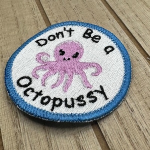 Può includere: Una toppa ricamata bianca con un bordo blu. La toppa presenta un polpo rosa con un viso sorridente e il testo "Don't Be a Octopussy".