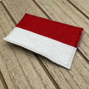 Op de afbeelding: Een rode en witte stoffen patch met een gestikte rand. De patch ligt op een houten oppervlak.