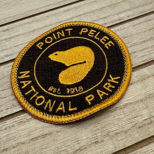 Könnte beinhalten: Ein runder, goldfarbener Aufnäher mit schwarzer Stickerei. Der Aufnäher zeigt den Text "Point Pelee National Park" mit einer Biber-Silhouette und dem Text "Est. 1918".