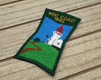 May include: A green and yellow embroidered patch with a lighthouse and a winding path. The text "WEST COAST TRAIL" is on the patch.