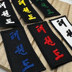 Peut inclure: Six patchs en tissu noir avec du texte brodé en blanc, jaune, vert, rouge et bleu. Le texte sur chaque patch est "跆拳道", qui est le mot coréen pour Taekwondo.