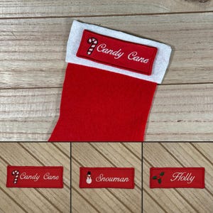 Könnte beinhalten: Drei rote Filz-Weihnachtsstrümpfe mit weißem Filzbesatz. Jeder Strumpf hat ein anderes gesticktes Design: ein Zuckerstange, ein Schneemann und Stechpalme. Die Strümpfe sind mit "Candy Cane", "Snowman" und "Holly" beschriftet.
