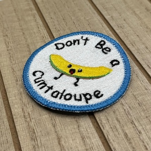 Può includere: Una toppa ricamata bianca con un bordo blu. La toppa presenta un melone cantalupo dei cartoni animati con un'espressione sorpresa e il testo "Don't Be A Cantaloupe".