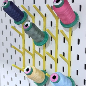 May include: A white pegboard with yellow thread spool holders, each holding a different color of thread. The thread colors include navy blue, pink, gray, light yellow, and light blue. The spools are green and pink.