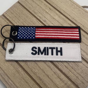 May include: Two rectangular keychains. One features the American flag with red and white stripes and a blue field of stars. The other is white with the name "SMITH" in black letters. Both have black metal key rings.
