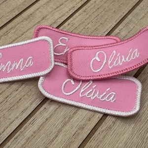 Op de afbeelding: Vier roze stoffen naamstickers met witte geborduurde letters. De namen zijn Emma, Olivia, Olivia en Emma.