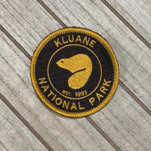 May include: Circular patch with a black central area and a gold border. The patch displays the text "KLUANE NATIONAL PARK" in gold lettering, with a gold bear silhouette in the centre. The text "est. 1993" is also included.