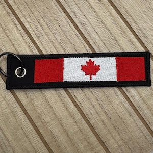 May include: A rectangular keychain featuring the Canadian flag. The flag design includes red bars on each end, a white center with a red maple leaf, and a black border. A metal ring is attached for keys.