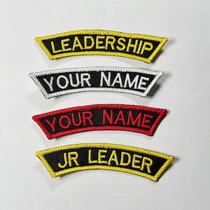 May include: Four embroidered patches with arched designs. The top patch reads "LEADERSHIP" in yellow on a black background. Below it, a patch with "YOUR NAME" in white on black. The third patch has "YOUR NAME" in red on black. The bottom patch says "JR LEADER" in yellow.