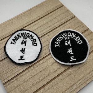 May include: Two round embroidered patches with the text "Taekwondo" in black lettering on a white background and a white background with black lettering. The patches have a black and white symbol in the center.