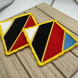 Op de afbeelding: Twee ruitvormige patches met gele stiksels. De patches zijn gemaakt van rood, zwart, wit en blauw stof. De patches liggen op een houten oppervlak.